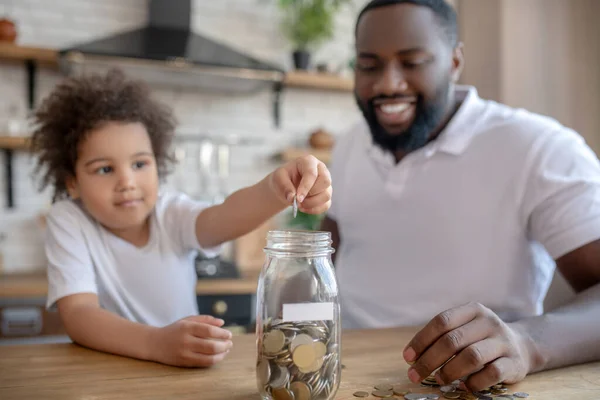 Cute curly-haired kid putting coins into the moneybank — Stock Photo, Image