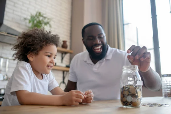 Dark-skinned bearded man putting coins into the moneybank — Stock Photo, Image