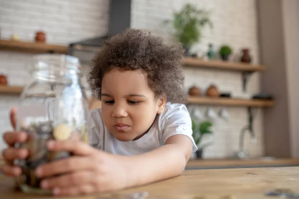 Lindo niño de pelo rizado en una camiseta blanca sosteniendo un banco de dinero y mirando insatisfecho — Foto de Stock