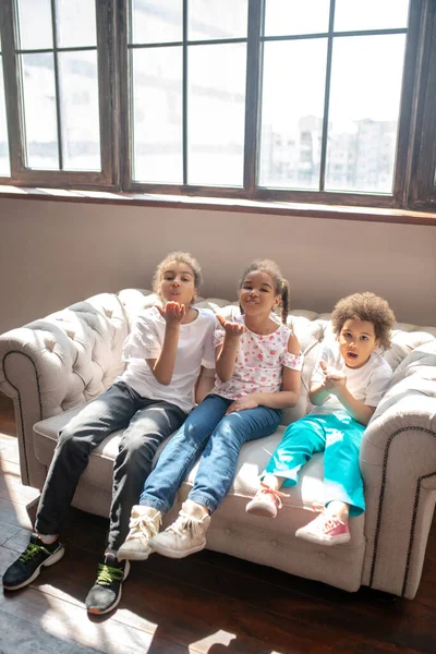 Drei lockige Kinder sitzen auf dem Sofa und versenden Luftküsse — Stockfoto