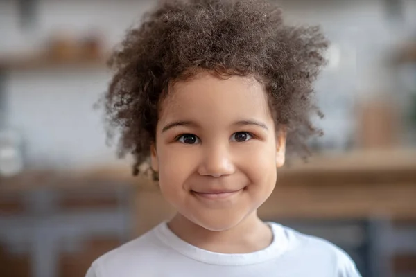 Beyaz tişörtlü tatlı kıvırcık saçlı çocuk pozitif gülümsüyor. — Stok fotoğraf