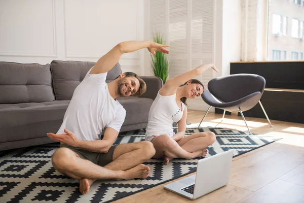 Pareja joven en camisetas blancas inclinadas mientras hacen ejercicio en línea juntos en casa —  Fotos de Stock