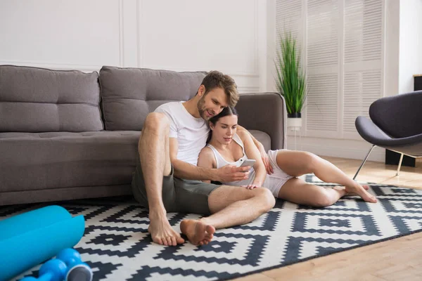 Pareja joven sentada en el suelo después del entrenamiento, viendo algo en línea y sintiéndose relajada — Foto de Stock