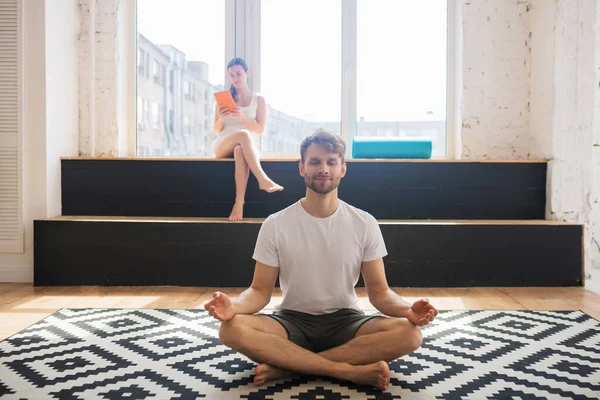 Bearded man in white tshirt sitting on the floor and meditating in lotus pose — Stock Photo, Image
