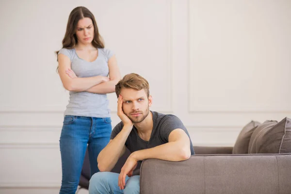 Mujer joven de pie con los brazos cerrados mientras su marido sentado con mirada deprimida — Foto de Stock