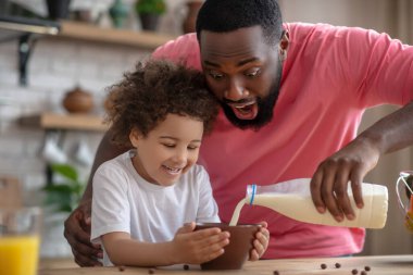 Afrikalı Amerikalı sakallı adam kızının tabağına süt döküyor.