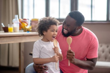 Kıvırcık saçlı şirin bir çocuk ve babası kaşık tutup şarkı söylüyor.