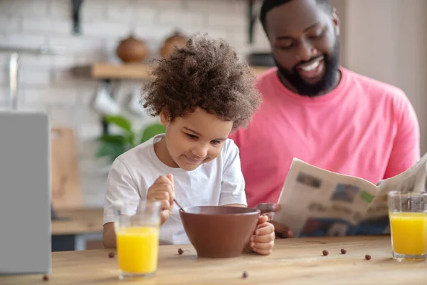 Sötét hajú afro-amerikai apa újságot olvas, miközben a gyereke reggelizik. — Stock Fotó