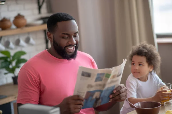 Dark-haired african american father reading a newspaper while having breakfast with his daughter — Stock Photo, Image