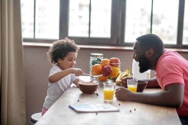 Hombre afroamericano en una camiseta rosa entreteniendo a su hija mientras ella come —  Fotos de Stock