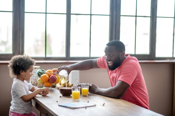 Hombre afroamericano con una camiseta rosa sentado a la mesa y desayunando con su hija —  Fotos de Stock