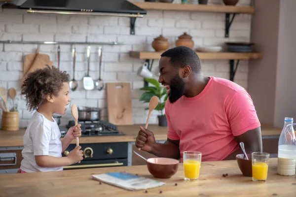 Lindo niño con el pelo rizado y su padre sosteniendo cucharas como micrófonos y cantando antes de desayunar —  Fotos de Stock