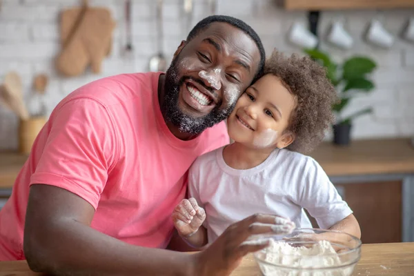 Σκούρο δέρμα πατέρας φαίνεται αστείο με το πρόσωπο λεκιασμένο με αλεύρι, η κόρη του χαμογελά — Φωτογραφία Αρχείου