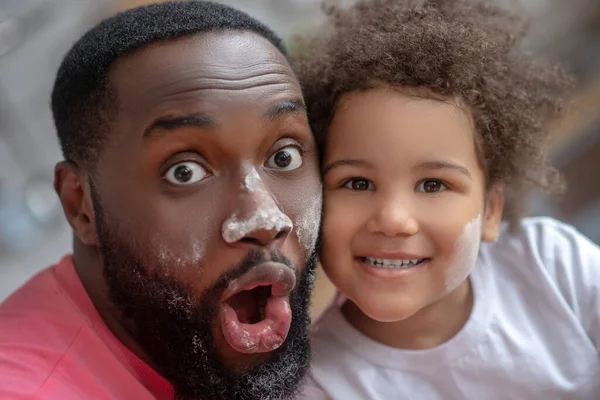 Dark-skinned father opening his mouth and looking funny with face stained with flour, his daughter smiling — Stock Photo, Image