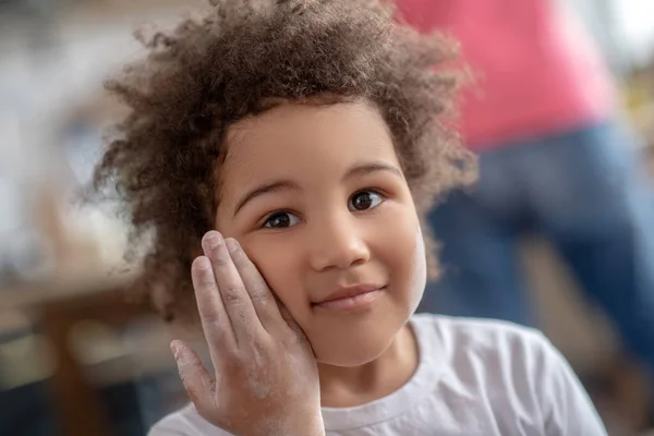 Hübsches lockiges Kind berührt ihre Wange mit einer mit Mehl befleckten Hand und sieht glücklich aus — Stockfoto