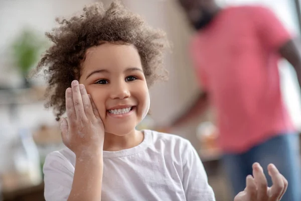 Pretty curly-haired kid touching her cheek with hand stained with flour — Stock Photo, Image