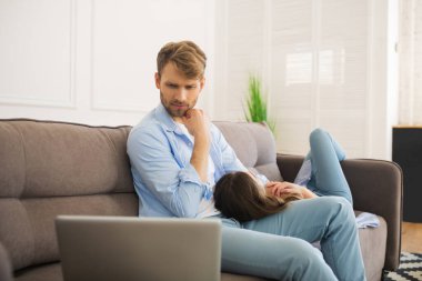 Genç adam dizüstü bilgisayarda çalışıyor ve ciddi görünüyor, karısı dinleniyor.