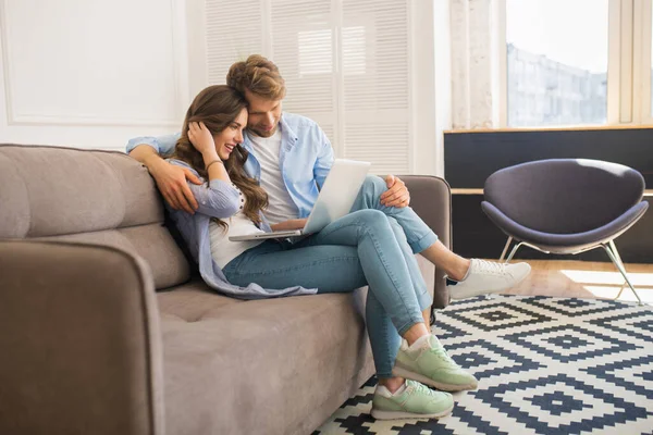 Pareja joven en denim sentada en un sofá viendo algo en línea y sintiéndose cómoda — Foto de Stock