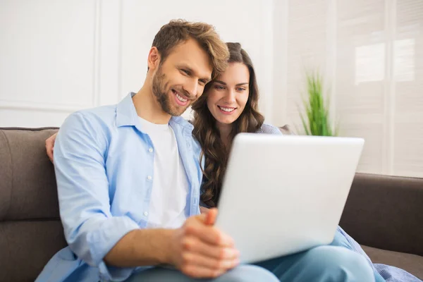 Молодая пара в джинсах сидит на диване и смотрит что-то онлайн и улыбается — стоковое фото