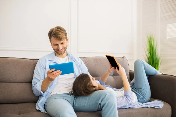 Sonriente joven mirando algo en la tableta, su esposa mintiendo y sosteniendo su dispositivo —  Fotos de Stock