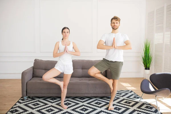 Tineri cuplu în tricouri albe face yoga la domiciliu în picioare în poză copac — Fotografie, imagine de stoc