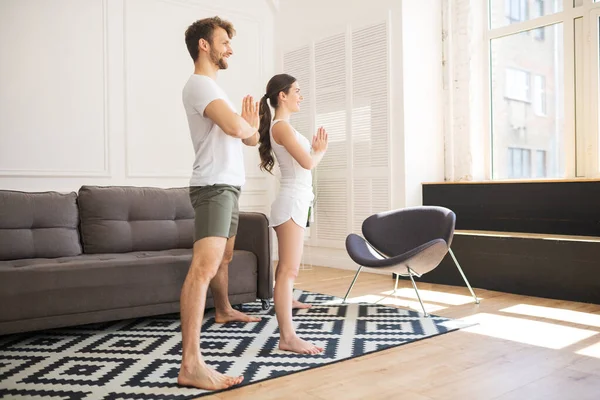 Pareja joven en camisetas blancas haciendo yoga en casa y buscando involucrado —  Fotos de Stock