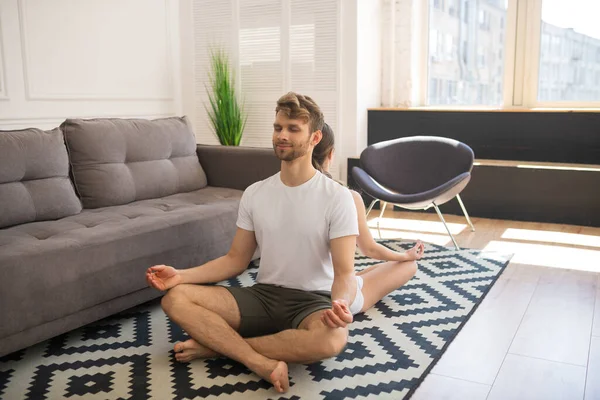 Young couple in white tshirts sitting in lotus pose back to back and looking calm — Stock Photo, Image