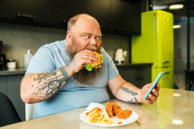 Sakallı şişman adam akıllı telefondan burger yiyor.