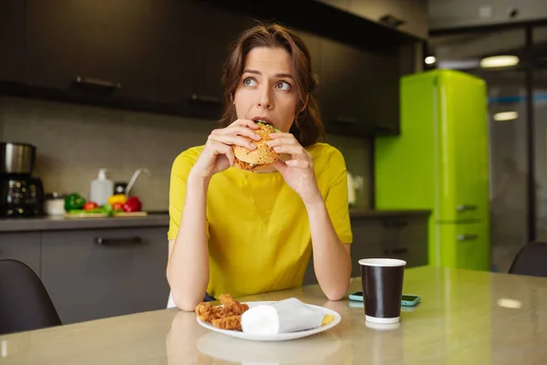 Sötét hajú lány leharap egy hamburgert, oldalra néz. — Stock Fotó