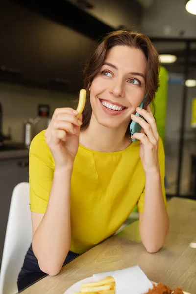 Милая улыбающаяся женщина держит картошку фри разговаривая со смартфоном — стоковое фото