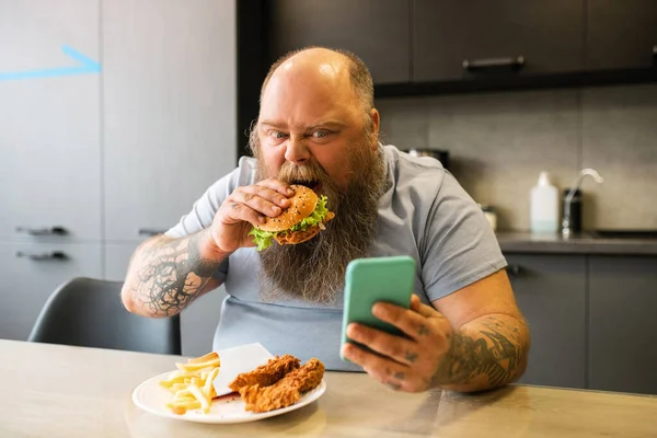 Barba tatuata uomo coinvolto con smartphone e hamburger . — Foto Stock