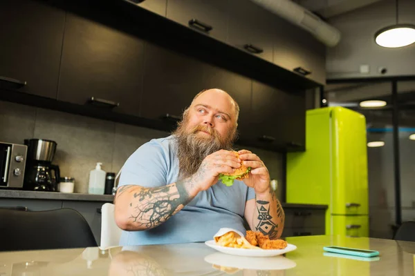 Szczęśliwy duży człowiek z hamburgerem w rękach przy stole — Zdjęcie stockowe