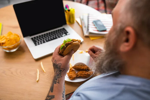 Uomo calvo seduto al tavolo con un hamburger in mano — Foto Stock