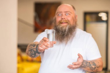 Kel sakallı tombul adam elinde bir bardak su tutuyor.