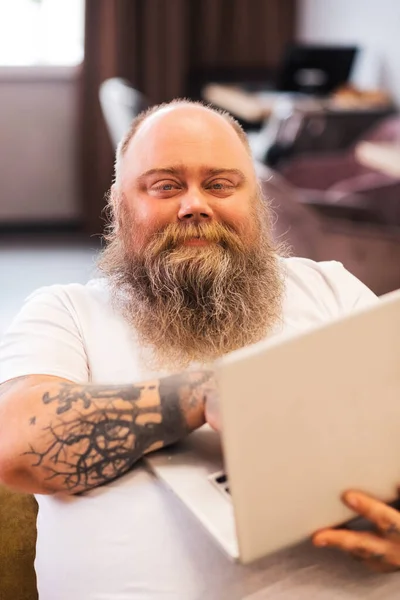 Bald bearded plump man sitting with a laptop in hands