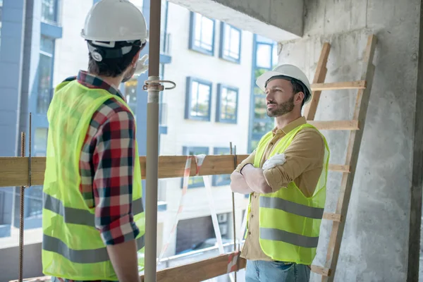 Trabajadores de la construcción con chalecos y guantes amarillos de pie cerca de la ventana, discutiendo algo —  Fotos de Stock