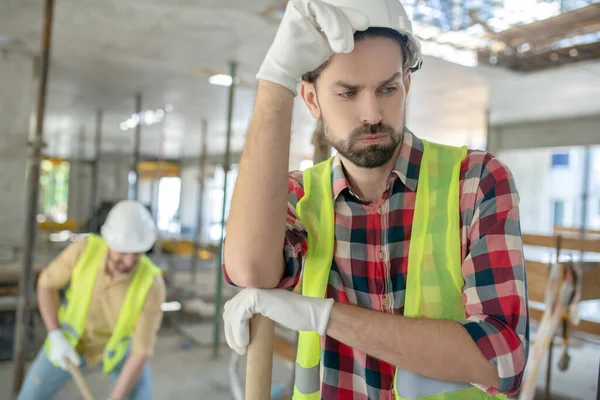 Trabajador de la construcción cansado en chaleco amarillo y guantes tocando su casco, suspirando, su compañero de trabajo trabajando golpeándolo —  Fotos de Stock