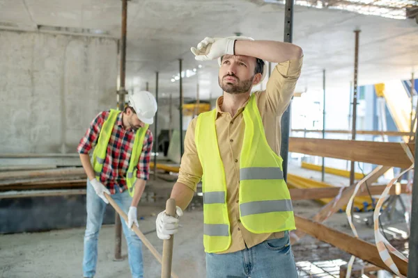 Tired building worker in yellow vest and gloves mopping his brow, his coworker working behing him — Stock Photo, Image