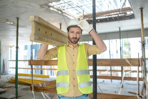 身穿黄色背心、戴着手套、肩上扛着木板、用手触摸头盔的建筑工人累坏了 — 图库照片