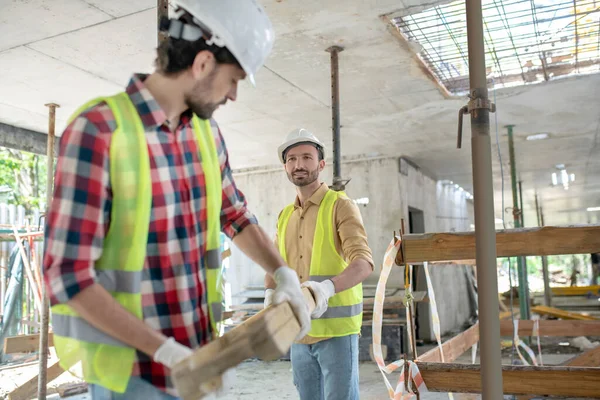 Building workers in yellow vests and gloves carrying together wooden board — Stock Photo, Image