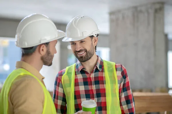 Trabajadores de la construcción en chalecos amarillos y cascos bebiendo café, hablando, sonriendo —  Fotos de Stock