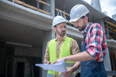 İşçiler vincin önünde duruyor, planları kontrol ediyor, beyin fırtınası yapıyorlar.