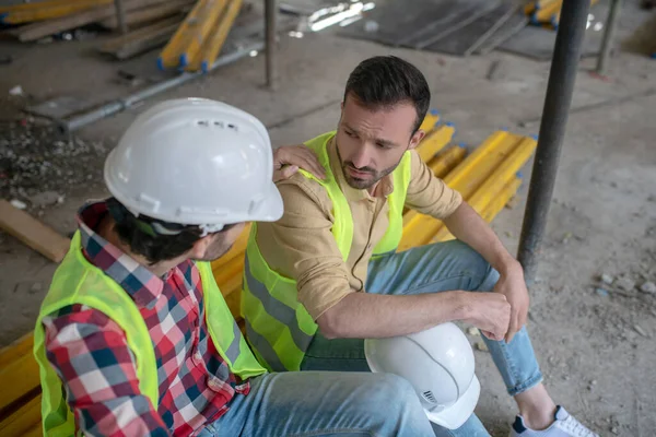 Trabajadores de la construcción cansados en chalecos amarillos sentados en las tablas, uno de los hombres poniendo su mano en el hombro de su compañero de trabajo — Foto de Stock