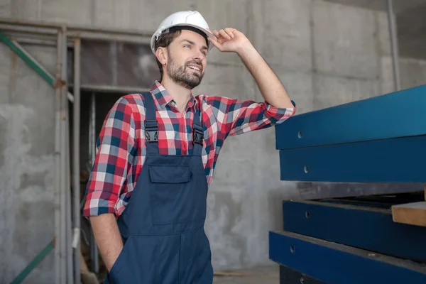 Building worker in helmet and checked shirt standing with one hand in pocket, touching his helmet with another — Stock Photo, Image