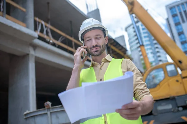 Trabalhador da construção em capacete e colete amarelo segurando planta, falando ao telefone — Fotografia de Stock