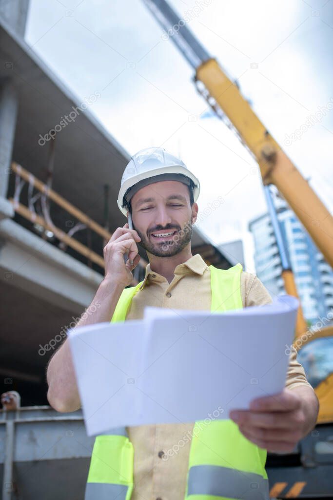 Building worker in helmet and yellow vest holding blueprint, talking on the phone, smiling