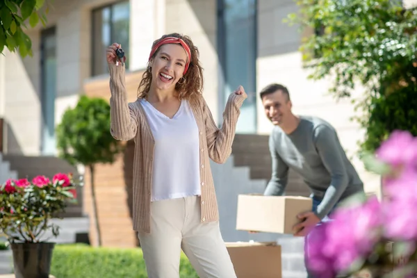 Mujer sonriente sosteniendo una llave de la nueva casa y sintiéndose genial, su marido de pie cerca de los cartones — Foto de Stock