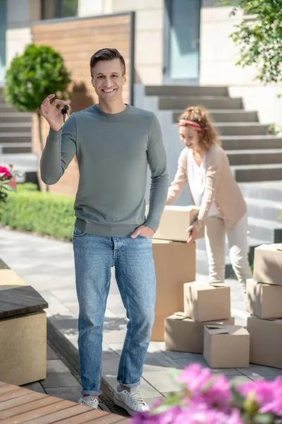 Sonriente hombre sosteniendo una llave de la nueva casa y sintiéndose feliz, su esposa de pie cerca de los cartones — Foto de Stock