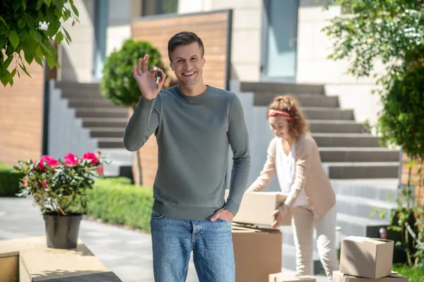 Sonriente hombre sosteniendo una llave de la nueva casa y sintiéndose muy bien, su esposa de pie cerca de los cartones — Foto de Stock