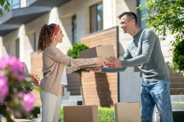 Mujer en pantalones blancos pasando un cartón a su marido mientras se muda a una casa nueva — Foto de Stock